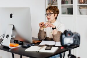 Foto de uma mulher sentada em frente a um computador e a uma câmera para ilustrar artigo sobre consultoria de recolocação profissional.